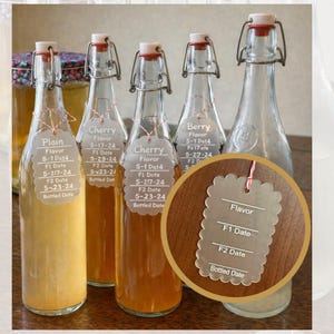 May include: A collection of glass bottles with swing-top lids, containing different coloured liquids. Each bottle has a tag indicating the flavour and dates. Flavours include "Plain", "Cherry", and "Berry". The bottles are on a wooden surface.