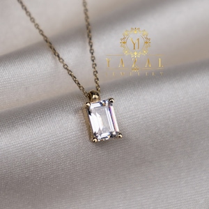 May include: A gold chain necklace with a clear emerald-cut gemstone pendant. The necklace is on a white fabric background.