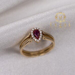 Puede incluir: Un anillo de oro con un rubí en forma de pera rodeado por un halo de pequeños diamantes. El anillo está sobre un fondo de satén blanco.