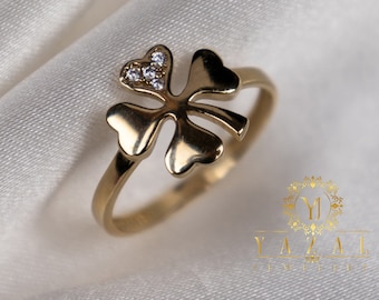 Puede incluir: Un anillo de oro con un diseño de trébol de cuatro hojas. El centro del trébol es una forma de corazón con pequeñas piedras transparentes.