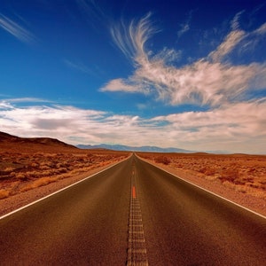 Könnte beinhalten: Eine lange, gerade Asphaltstraße erstreckt sich unter einem strahlend blauen Himmel mit federartigen Wolken in die Ferne. Die Straße wird von einer trockenen, rötlich-braunen Wüstenlandschaft flankiert, mit fernen Bergen am Horizont.