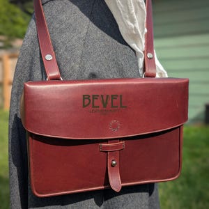 Puede incluir: Bandolera de cuero burdeos con cierre de solapa y correa ajustable para el hombro. La bolsa presenta el texto "BEVEL - LEATHERCRAFT" en la parte delantera. La bolsa es rectangular y tiene un cierre de hebilla.