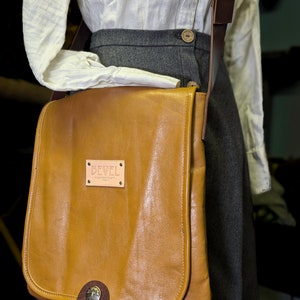May include: A tan leather messenger bag with a brown strap and a metal buckle. The bag has a small metal tag that says "BEVEL" on it.
