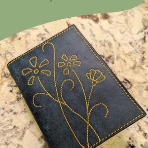 Peut inclure: Un petit portefeuille de carte en cuir noir avec un motif floral cousu en fil jaune. Le portefeuille est sur une surface brun clair. "BEVEL LEATHERCRAFT." est imprimé dans le coin supérieur droit de l'image.
