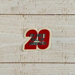 May include: A sticker with a red number 20 shape and the text "THE GREAT WALL OF MINNESOTA" in white and green. The sticker is on a white wooden surface.
