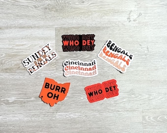 May include: A collection of six stickers featuring the Cincinnati Bengals logo and the phrase "Who Dey". The stickers are in various shades of orange and black. The stickers are on a light wood background.