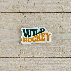 May include: A white sticker with the words "WILD HOCKEY" in dark green and orange block letters. The sticker has a retro design and is placed on a light-colored, textured wooden surface.