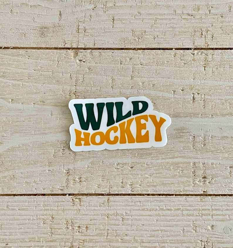 May include: A white sticker with the words "WILD HOCKEY" in dark green and orange block letters. The sticker has a retro design and is placed on a light-colored, textured wooden surface.