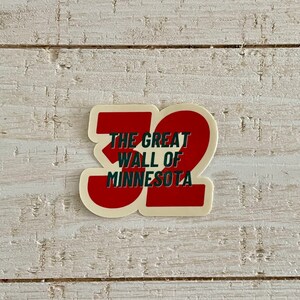May include: A sticker with a red and white design featuring the text "THE GREAT WALL OF MINNESOTA" in green and white, with the number "32" in red. The sticker is on a white wooden surface.