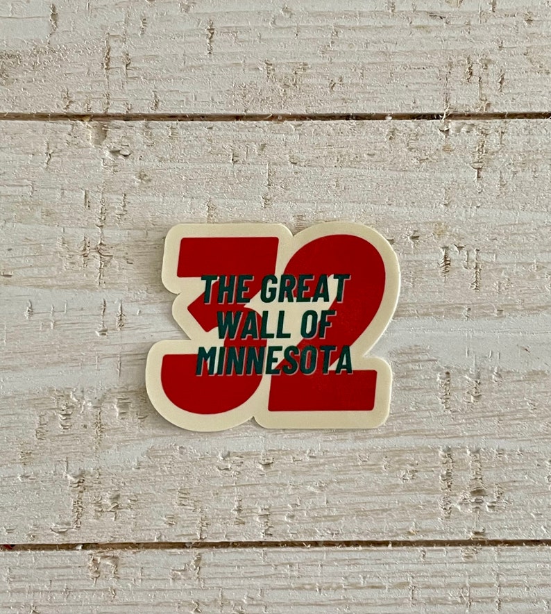 May include: A sticker with a red and white design featuring the text "THE GREAT WALL OF MINNESOTA" in green and white, with the number "32" in red. The sticker is on a white wooden surface.