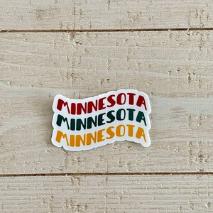 May include: A white sticker with the word "MINNESOTA" stacked three times in red, green, and yellow. The sticker is on a light wood-grain surface.