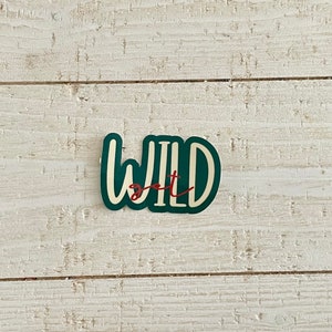 May include: A sticker with the words "WILD" in white, outlined in dark green, with the word "get" in red script. The sticker is on a white, weathered wood surface.