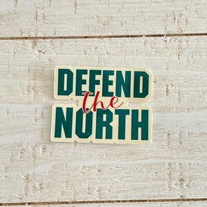 May include: A sticker with the words "DEFEND the NORTH" in dark green block letters. The word "the" is in red script. The sticker has a cream-colored background and is set against a white wooden surface.