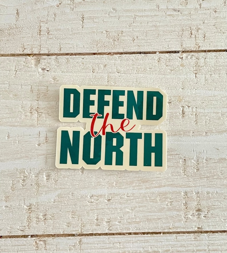 May include: A sticker with the words "DEFEND the NORTH" in dark green block letters. The word "the" is in red script. The sticker has a cream-colored background and is set against a white wooden surface.