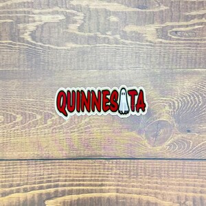 May include: A sticker with the word "QUINNESATA" in red, outlined in white, with a small ghost graphic. The sticker is on a wooden surface.