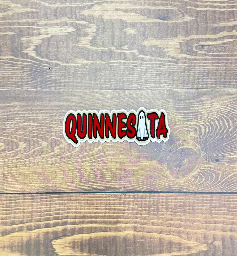 May include: A sticker with the word "QUINNESATA" in red, outlined in white, with a small ghost graphic. The sticker is on a wooden surface.