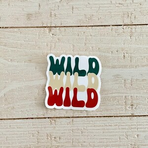 May include: A sticker with the word "WILD" stacked three times in different colors: dark green, tan, and red. The sticker is on a light wood surface.