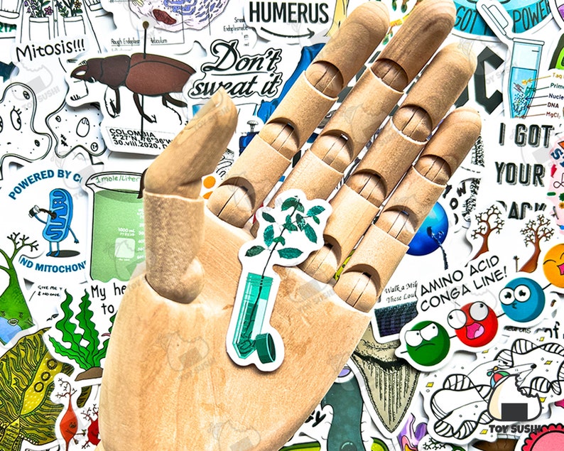 Puede incluir: Una mano de artista de madera est&aacute; rodeada de varias pegatinas. Una pegatina presenta una planta en un tubo de ensayo. Otras pegatinas tienen texto como "Mitosis!!!", "Don't sweat it" y "Amino Acid Conga Line!"