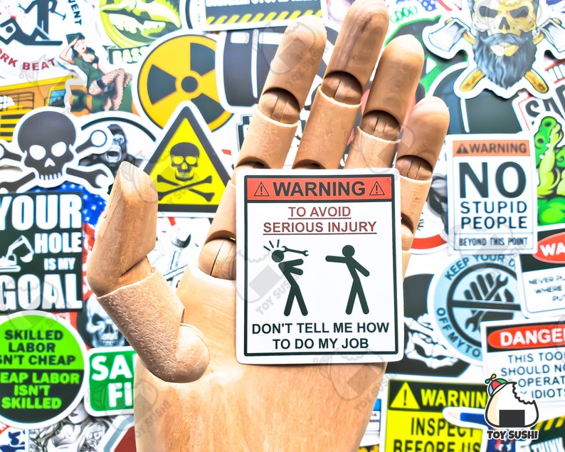 May include: A white warning sticker with black text and a black and white illustration. The sticker reads "Warning: To avoid serious injury, don't tell me how to do my job". The illustration shows a stick figure holding a wrench and hitting another stick figure on the head.