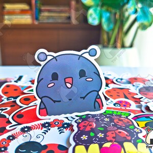 May include: A collection of colorful ladybug stickers with a cartoon ladybug sticker in the center. The cartoon ladybug is blue and gray with a big smile and two antennas. The stickers are perfect for decorating journals, laptops, water bottles, and other items.