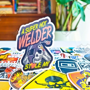 May include: A sticker with the text "A SUPER HOT WELDER STOLE MY" in pink and orange. The sticker features a skull wearing a welding helmet with a flame graphic. The sticker is surrounded by other stickers.