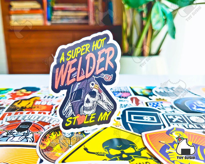 May include: A sticker with the text "A SUPER HOT WELDER STOLE MY" in pink and orange. The sticker features a skull wearing a welding helmet with a flame graphic. The sticker is surrounded by other stickers.