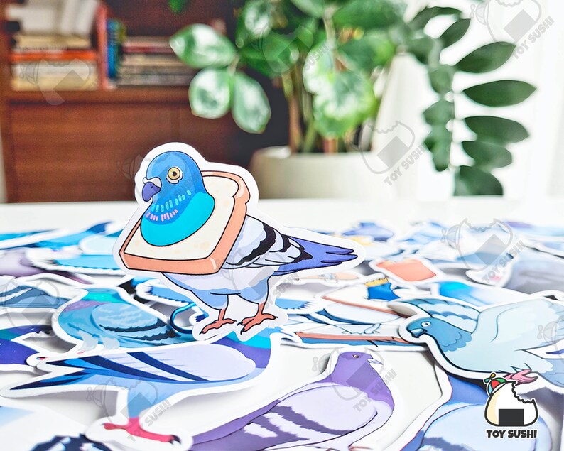 May include: A sticker sheet featuring a cartoon pigeon wearing a slice of bread on its head. The sticker sheet also includes other pigeon designs in various poses and colours. The sticker sheet is on a white surface with a plant in the background.