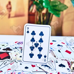 May include: A white playing card with a black nine of clubs symbol. The card is surrounded by other playing cards on a white surface.