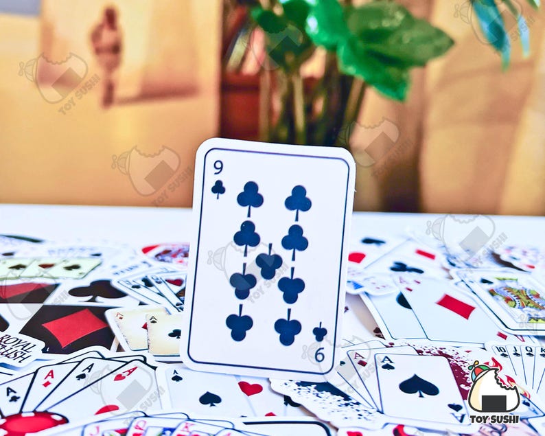 May include: A white playing card with a black nine of clubs symbol. The card is surrounded by other playing cards on a white surface.