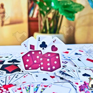 May include: A set of red dice with white dots, surrounded by playing cards. The dice are in the shape of a cube and have a heart symbol on one side. The cards are all face down and are arranged in a random pattern.