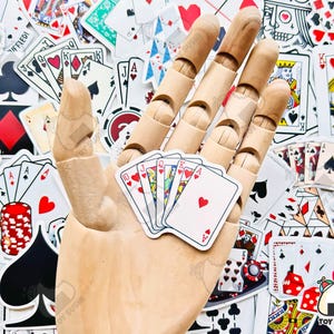 May include: A wooden hand holding a set of playing cards. The cards are all face up and show the numbers 10, Jack, Queen, King, and Ace. The cards are all hearts. The hand is surrounded by a variety of playing cards, including a royal flush, a joker, and a few other cards.