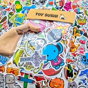 May include: A clear plastic bag filled with colorful stickers. The stickers feature various designs, including a bomb, a bunny, a cross, a pumpkin, and a cartoon character. The bag is labeled "Toy Sushi".