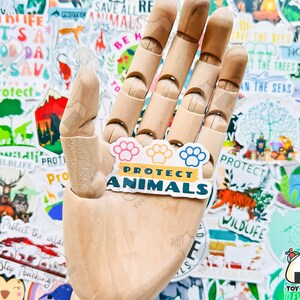 May include: A sticker with a paw print design and the text "Protect Animals" in yellow and blue letters on a white background. The sticker is held by a wooden hand.