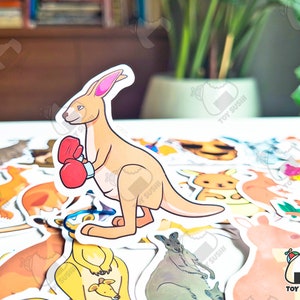 May include: A sticker of a cartoon kangaroo wearing boxing gloves. The kangaroo is brown and has a white belly. The sticker is on a white background with other stickers of animals.