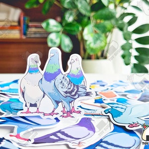 May include: Three colourful pigeon stickers with blue, green and white accents. The stickers are on a white surface with other stickers in the background.