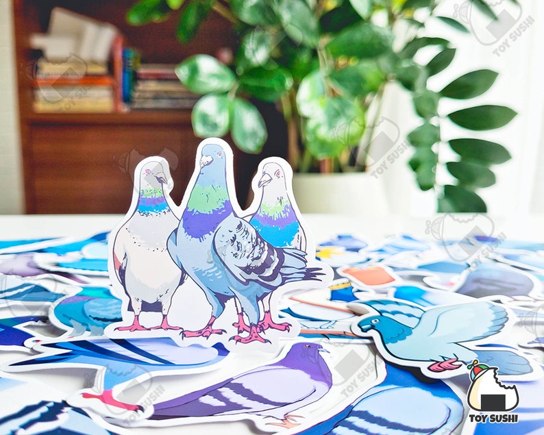 May include: Three colourful pigeon stickers with blue, green and white accents. The stickers are on a white surface with other stickers in the background.
