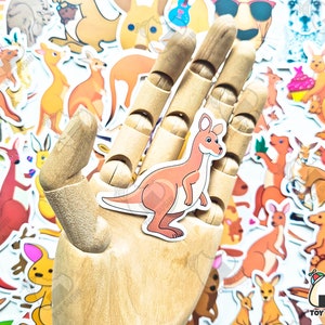May include: A wooden hand holding a sticker of a brown kangaroo with a white belly. The sticker is surrounded by other stickers of kangaroos, some with a cupcake, some with sunglasses, and some with a guitar. The stickers are on a white background.