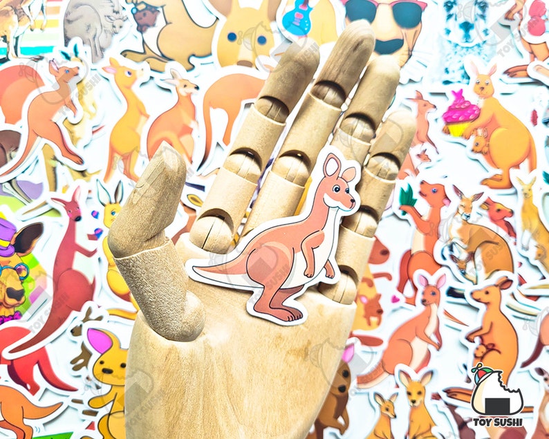 May include: A wooden hand holding a sticker of a brown kangaroo with a white belly. The sticker is surrounded by other stickers of kangaroos, some with a cupcake, some with sunglasses, and some with a guitar. The stickers are on a white background.