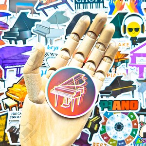 May include: A hand holding a sticker of a colorful grand piano with a black outline. The sticker is surrounded by other stickers of pianos, musical notes, and other music-related designs.