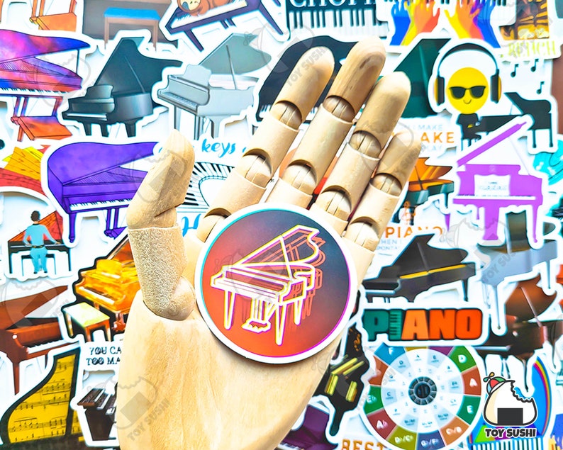 May include: A hand holding a sticker of a colorful grand piano with a black outline. The sticker is surrounded by other stickers of pianos, musical notes, and other music-related designs.