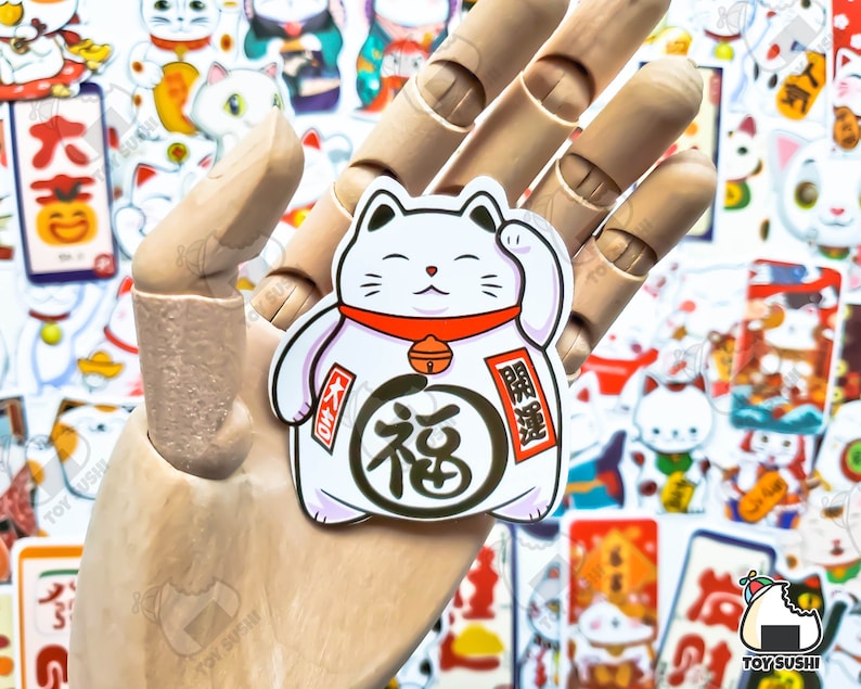 May include: A white lucky cat figurine with a red collar and a gold bell. The cat is holding up its right paw and has a happy expression on its face. The figurine is surrounded by other Japanese-themed stickers.