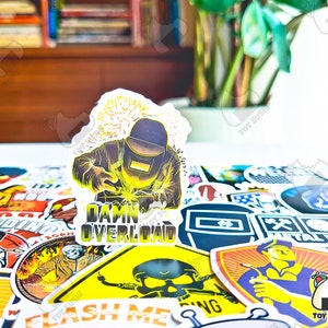 May include: A collection of stickers with various designs, including a sticker featuring a welder with the text "Damn Overload." Other stickers depict welding-related imagery and text. The stickers are colorful and have a glossy finish.