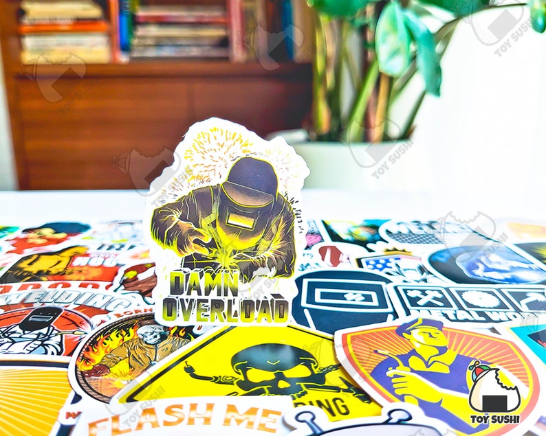 May include: A collection of stickers with various designs, including a sticker featuring a welder with the text "Damn Overload." Other stickers depict welding-related imagery and text. The stickers are colorful and have a glossy finish.
