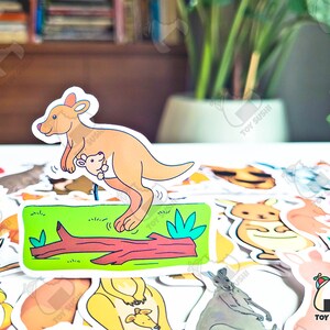 May include: A colorful sticker of a kangaroo with a joey in its pouch. The kangaroo is standing on a log with green grass and a leaf. The sticker is part of a set of animal stickers.
