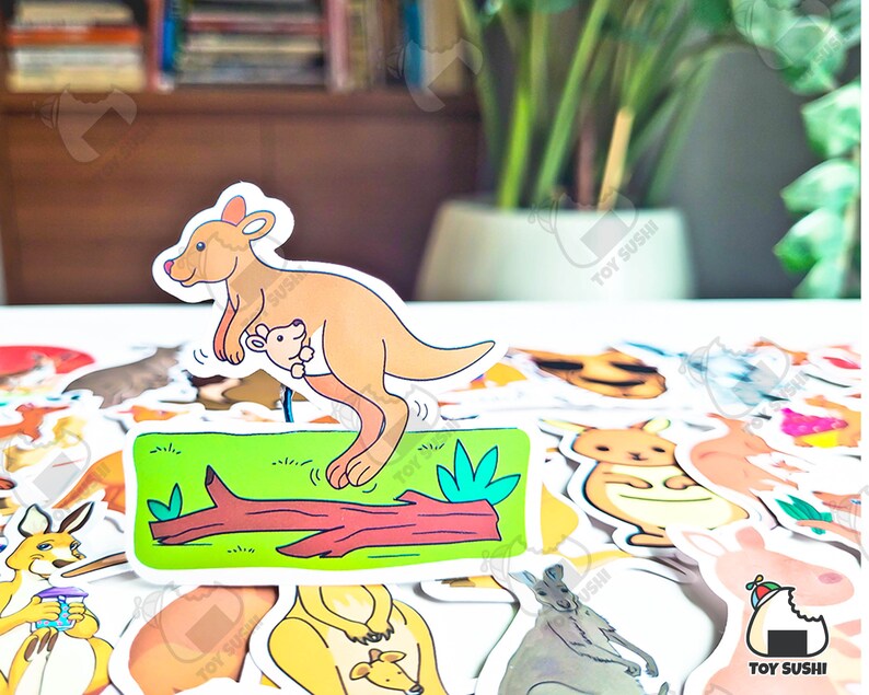 May include: A colorful sticker of a kangaroo with a joey in its pouch. The kangaroo is standing on a log with green grass and a leaf. The sticker is part of a set of animal stickers.