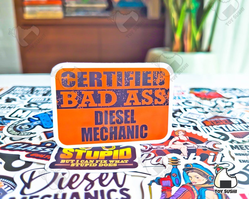 May include: A collection of colorful stickers with various designs, including a sticker that reads "Certified Bad Ass Diesel Mechanic".