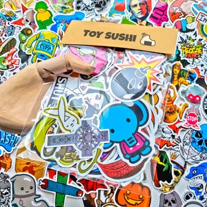May include: A clear plastic bag filled with colorful stickers. The stickers feature various designs, including a bomb, a bunny, a cross, a pumpkin, and a cartoon character. The bag is labeled "Toy Sushi".