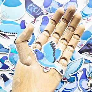 May include: A wooden hand holding a blue and white pigeon sticker. The sticker is surrounded by other pigeon stickers in various colours and designs.