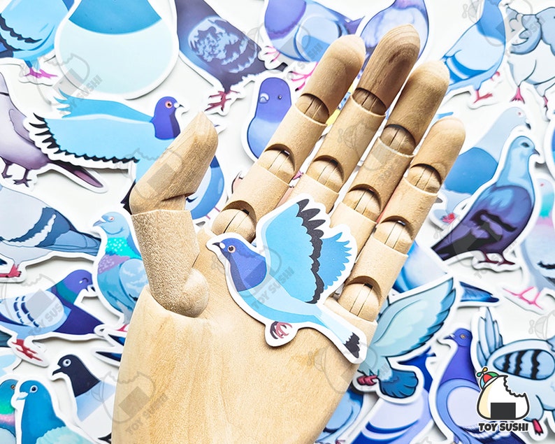 May include: A wooden hand holding a blue and white pigeon sticker. The sticker is surrounded by other pigeon stickers in various colours and designs.