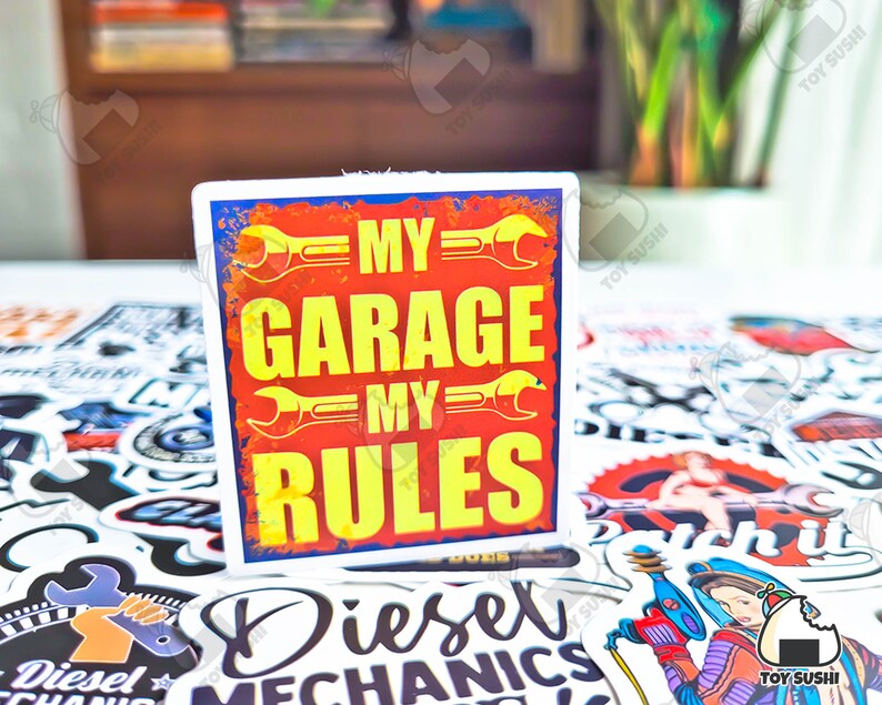 May include: A white sticker with a red and yellow design. The sticker says "My Garage My Rules" with a wrench graphic on each side of the text.